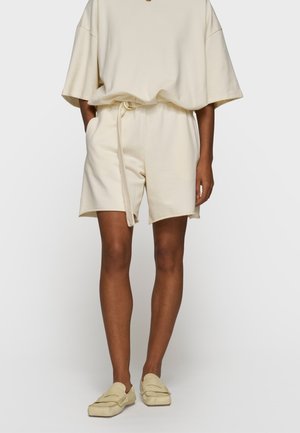 Persona che indossa pantaloncini beige con cintura in tessuto, top oversize a maniche corte abbinato e mocassini slip-on beige, in piedi con le mani nelle tasche.