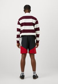 Μαρούν και λευκή ριγέ μπλούζα με μακριά μανίκια, μαύρα σορτς με κόκκινες λεπτομέρειες, μαύρα αθλητικά παπούτσια και ριγέ κάλτσες. Ορατές ραφές και λεπτομέρειες υφής.