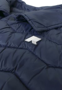 Giacca a vento blu marina con texture trapuntata, caratterizzata da un ampio patch con logo bianco. Finitura liscia e lucida con dettaglio di chiusura a pressione.