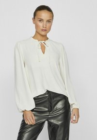 Blusa bianca a maniche lunghe con scollatura a chiave e dettagli con fiocco, abbinata a pantaloni in pelle nera lucida. Tessuto morbido con vestibilità rilassata.
