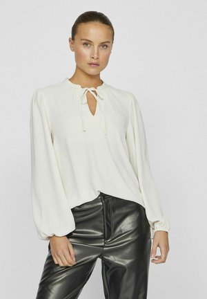 Blouse blanche à manches longues avec un col en forme de trou de serrure et des détails de nœud, associée à un pantalon en cuir noir brillant. Tissu doux avec une coupe décontractée.