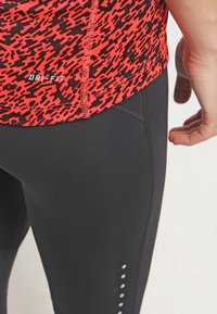Punainen ja musta kuviollinen urheilu-toppi, jossa on Dri-FIT -merkki; yhdistettynä mustiin legginsseihin, joissa on sivusauma ja valkoisia pistekoristeita.