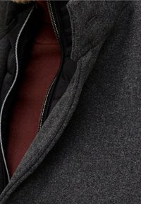 Cappotto grigio in misto lana con fodera interna nera trapuntata. Presenta un colletto alto e dettagli con zip a vista. Strato interno color ruggine visibile.