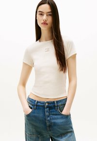 Beżowa dopasowana koszulka z bawełny z okrągłym dekoltem i krótkimi rękawami. Posiada małe, subtelne logo na piersi. Noszona z niebieskimi dżinsami.