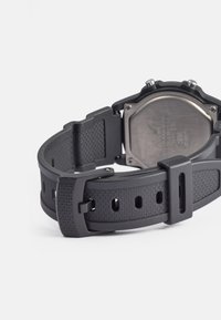 Relógio digital preto com uma bracelete de borracha texturizada, apresentando um fundo em aço inoxidável e fivela ajustável. Os detalhes notáveis incluem padrões retangulares.