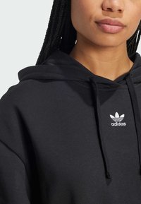 Čierna mikina s kapucňou, vyrobená z jemného materiálu, s prednou kangaroo vreckom, stiahnuteľnou kapucňou a bielym logom Adidas na hrudi.