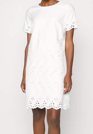 Robe blanche à manches courtes avec broderie ajourée sur les manches, l'ourlet et la moitié inférieure, portée par une personne debout devant un fond uni.