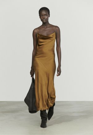 Woman wearing a brown satin slip dress and black boots, carrying a large black handbag, walking on a plain light gray floor and background.