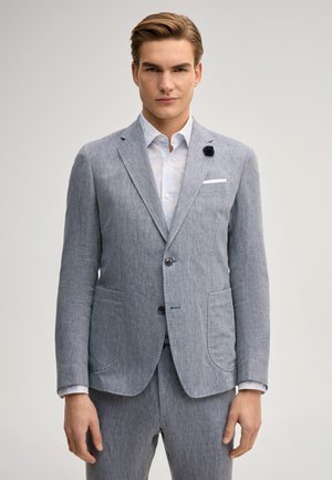 Junger Mann in einem hellgrauen, strukturierten Anzug mit weißem gemustertem Hemd und dunklem Reversanstecker, stehend vor einem einfarbigen Hintergrund.