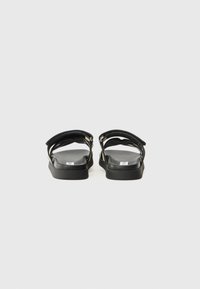 Black sandals with flat soles, featuring two wide straps and gold-tone hardware accents. Smooth texture and minimalist design.