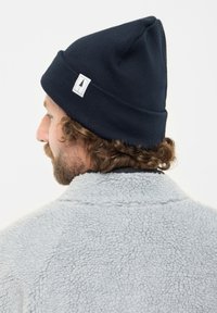 Uomo con capelli ricci e barba che indossa un berretto di maglia nero con un logo ad albero e una giacca in pile grigia chiara, di profilo.