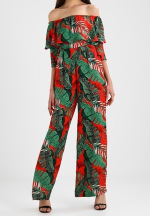 Kvinde iført rød off-shoulder jumpsuit med grøn tropisk bladprint og sorte åbne hæle, stående mod hvid baggrund.
