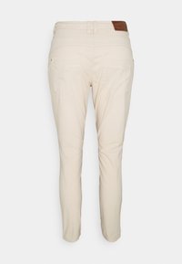 Béžové slim-fit kalhoty s dvěma zadními kapsami a hnědou koženou nášivkou na pase, vystavené na bílém pozadí.
