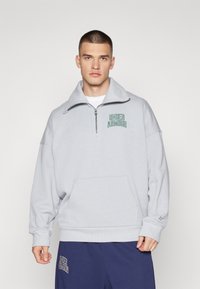 Sudadera de felpa gris con cuello alto y media cremallera, que cuenta con un bolsillo tipo canguro y el logo verde de "Under Armour" en el pecho.
