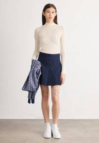 Top de gola alta em creme com textura canelada, combinado com uma saia plissada azul-marinho. A modelo segura uma jaqueta azul com padrão e usa ténis brancos.
