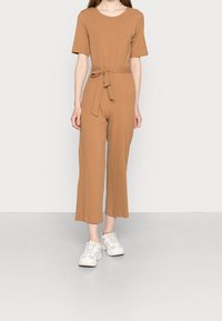 Brun, ribbade jumpsuit med rund halsringning, korta ärmar och midjeband med självknut. Byxorna är kapade och raka, i kombination med vita sneakers.