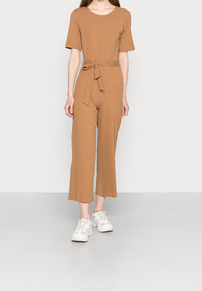 Brun, ribbade jumpsuit med rund halsringning, korta ärmar och midjeband med självknut. Byxorna är kapade och raka, i kombination med vita sneakers.