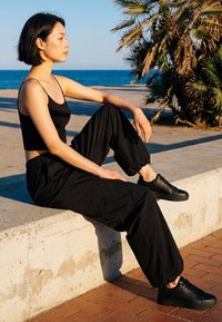 Young woman in black tank top and pants sitting on a concrete ledge by the sea, with palm trees in the background.