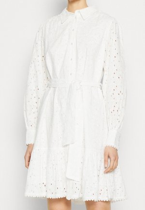 Femme portant une robe en dentelle ajourée blanche avec de longues manches bouffantes, boutonnée devant, col et ceinture à nouer à la taille, debout devant un fond uni.