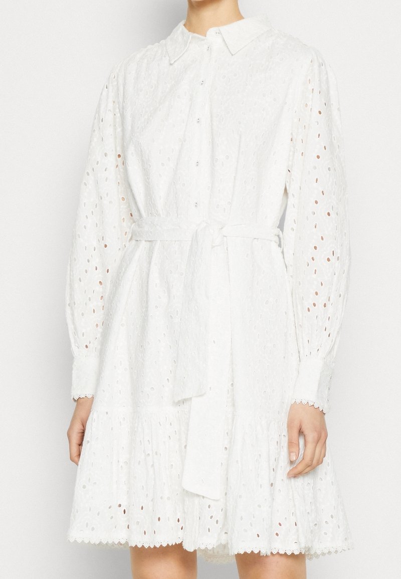 Femme portant une robe en dentelle ajourée blanche avec de longues manches bouffantes, boutonnée devant, col et ceinture à nouer à la taille, debout devant un fond uni.