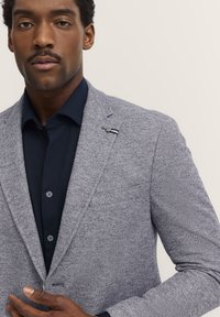 Grauer strukturierter Blazer mit subtiler Fischgrätmusterung, einem einzigen Knopf, Reverskragen und einem kontrastierenden dunkelblauen Hemd darunter.