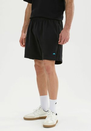 Hombre con pantalones cortos negros de cintura elástica, calcetines blancos y zapatillas blancas bajas, de pie sobre un fondo blanco.