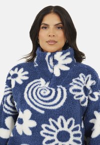 Donna con capelli scuri che indossa un pullover in pile blu con motivi floreali e spirali bianchi e un colletto alto con zip.