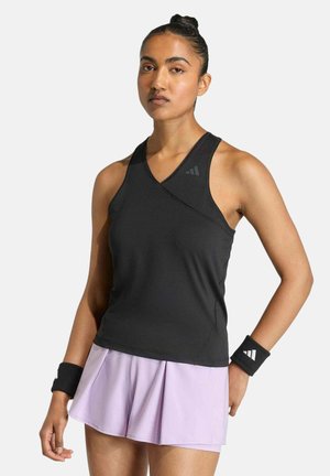 Mujer en forma con moño, vistiendo una camiseta deportiva sin mangas negra, falda plisada color púrpura claro y muñequeras negras, parada con la mano en la cadera.
