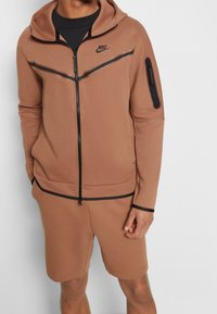 Hnědá souprava obsahující mikinu a šortky značky Nike. Mikina má plný zip, černé akcenty, kapuci se šnúrkou a kapsu na zip na rukávu.