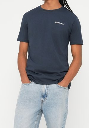 Persoon draagt een donkerblauw Replay T-shirt en lichtblauwe spijkerbroek met handen in de zakken, tegen een effen achtergrond.