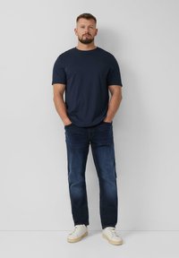 T-shirt in cotone blu navy con maniche corte, abbinata a jeans blu scuro e scarpe da ginnastica bianche. Il modello sta in piedi con le mani nelle tasche su uno sfondo chiaro.
