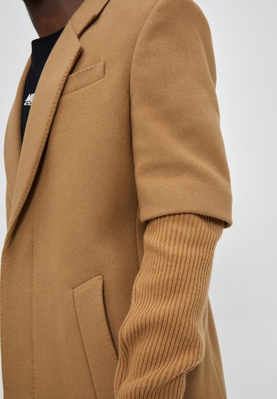 Camel-colored wool blend coat with a shawl collar, side pockets, and a ribbed knit sleeve accent. Smooth texture and tailored fit.
