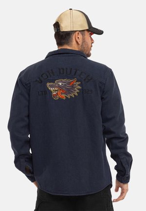 Homme portant une veste bleu marine avec une tête de loup brodée et le texte "Von Dutch LTD 1929" au dos, ainsi qu'une casquette de baseball beige et noire.