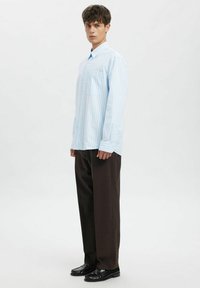 Lysblå stripete skjorte med lange ermer og knappelukking i kragen, kombinert med brune bukser og svarte sko. Merkbare detaljer inkluderer brystlomme.