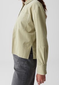 Personne portant une chemise vert pâle à manches longues avec une poche poitrine et un jean gris foncé, vue de côté sur un fond uni.