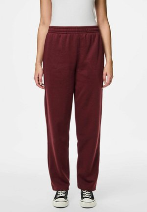 Pantalones de chándal burdeos con cinturilla elástica, piernas rectas y bolsillos laterales. Combinados con zapatillas negras con suelas blancas. Textura suave.