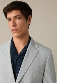 Helehall triibuline bleiser notslapiga, mida on kombineeritud tumesinise mustrilise särgiga. Sile kangastekstuur, istuv lõige ja peened detailid.