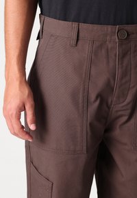 Pantalon cargo marron au design structuré, avec des poches avant, une couture solide et une fermeture à bouton. Le tissu a une texture lisse.