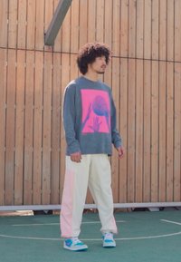 Grå hettegenser med en rosa basketballgrafikk, hvite og pastellrosa bukser, og blå sneakers. Står på en basketballbane med treverk som bakgrunn.