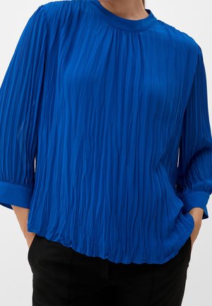 Femme portant un chemisier plissé bleu vif à manches trois-quarts et un pantalon noir, les mains partiellement dans les poches.