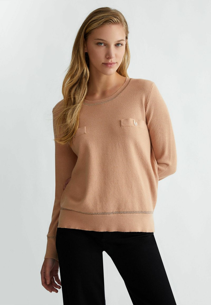 Maglione in maglia con texture pesca, dotato di due tasche frontali, scollatura rifinita e orlo a contrasto. Abbinato a pantaloni neri.