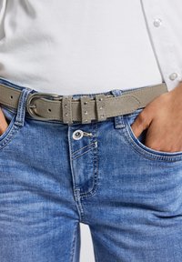 Hellblaue Jeans aus Denim mit einem grauen Wildledergürtel, der dekorative Nieten aufweist, getragen mit einem schlichten weißen Oberteil, die Hände in den Taschen.