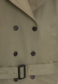 Trench-coat beige avec des boutons noirs croisés, une ceinture assortie avec une boucle rectangulaire noire et une poche poitrine côté droit.