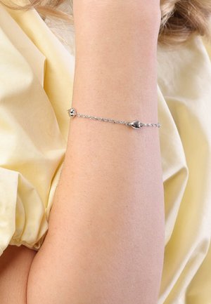 Silberne Kettenarmband mit herzförmigen Akzenten, das ein zartes Design und eine glatte Textur aufweist, getragen an einem Arm vor einem hellgelben Hintergrund.