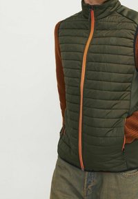 Zielona ocieplana kamizelka z pomarańczowym zamkiem błyskawicznym, z brązowymi żeberkowymi panelami bocznymi i wysokim kołnierzem. W zestawie kilka kieszeni.