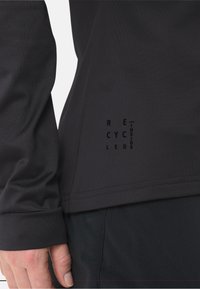 Top nero in tessuto a maniche lunghe con una texture liscia. Presenta un'etichetta di materiale riciclato stampata in nero vicino all'orlo.