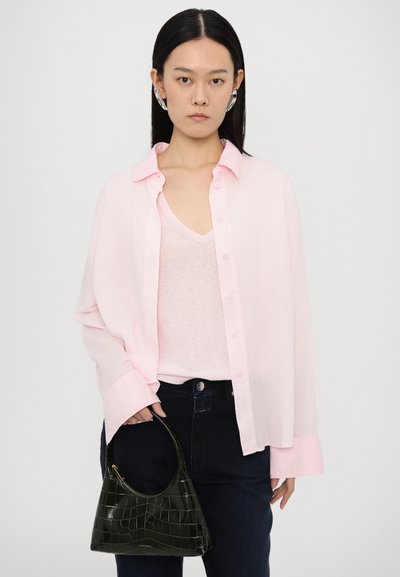 Chemise rose clair à manches longues sur un débardeur rose clair, associée à un jean bleu foncé. Tient un petit sac à main noir à motif crocodile.