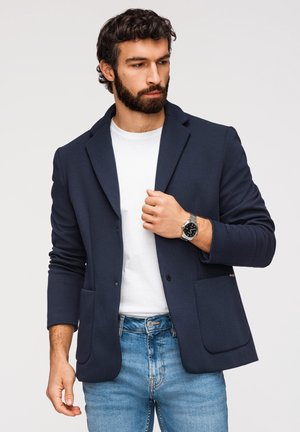 Homme portant un blazer bleu marine, un t-shirt blanc, un jean bleu clair et une montre-bracelet argentée, regardant vers le bas et ajustant le revers du blazer.