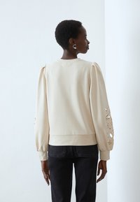 Felpa beige con maniche lunghe a sbuffo, caratterizzata da motivi floreali tagliati al laser sulle braccia e un orlo a coste. Indossata con jeans neri.