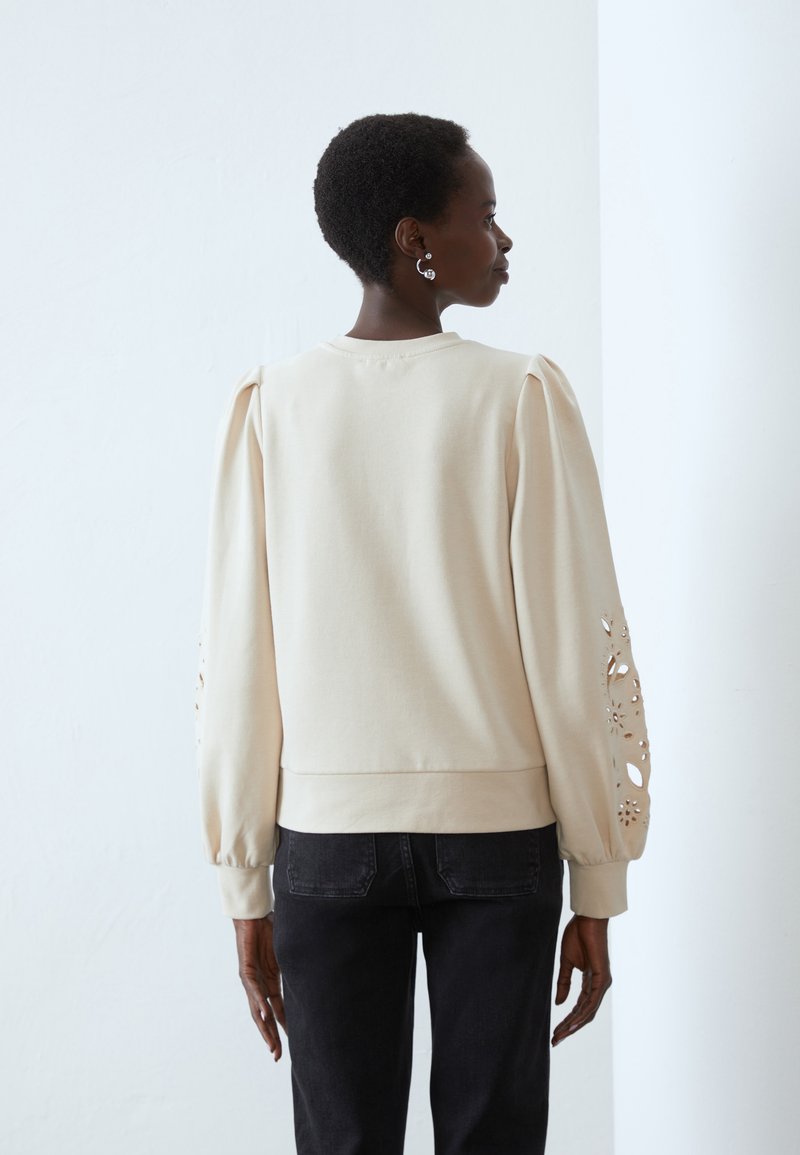 Felpa beige con maniche lunghe a sbuffo, caratterizzata da motivi floreali tagliati al laser sulle braccia e un orlo a coste. Indossata con jeans neri.